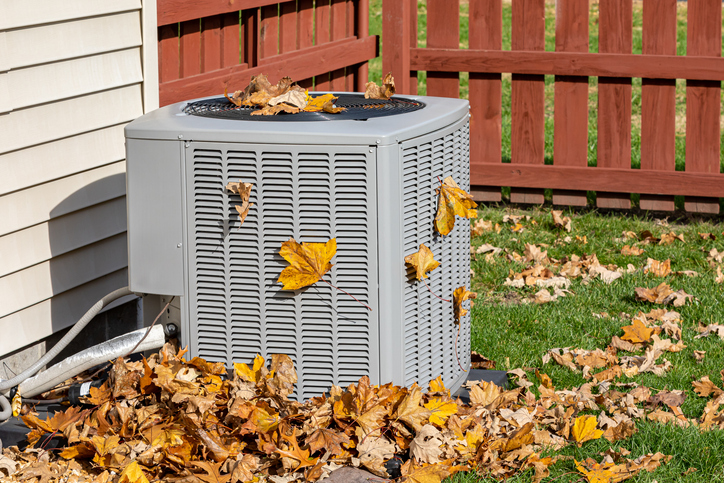 An air conditioning unit in a backyard, surrounded by fallen autumn leaves.